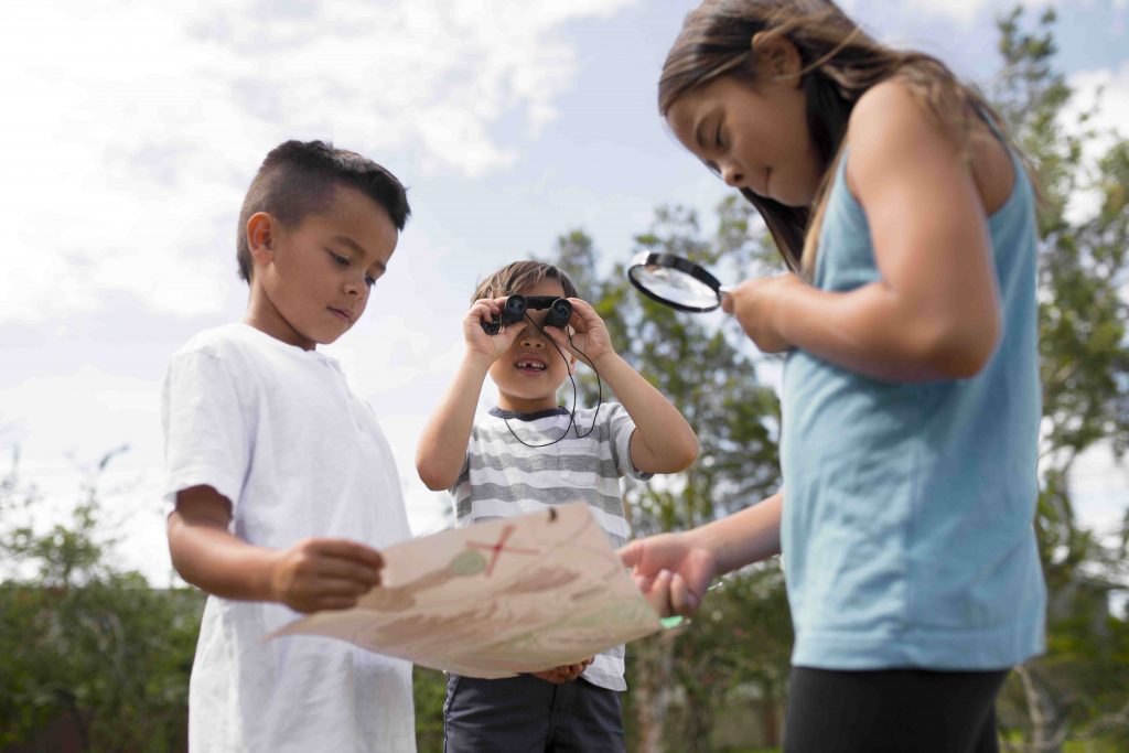 Três crianças ao ar livre, uma está segurando um binóculos, outra segura um mapa e a terceira segura uma lupa.