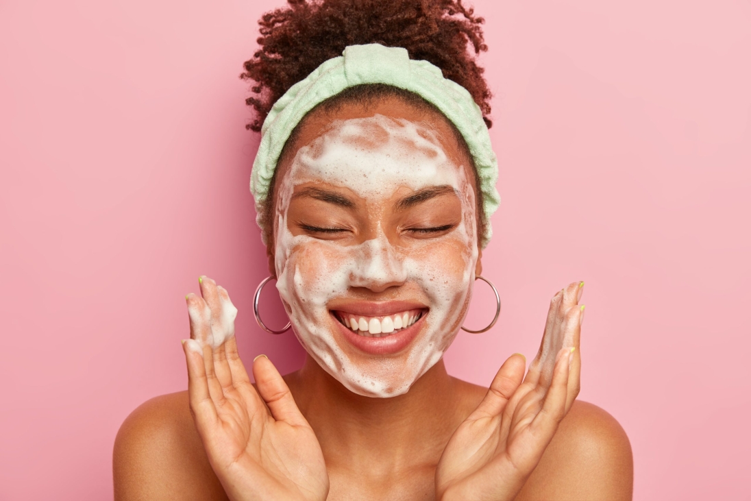 young-woman-raises-palms-over-face-keeps-eyes-closed-shows-white-teeth-uses-cleansing-foam-for-skin-care-gets-real-pleasure