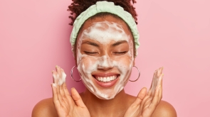 young-woman-raises-palms-over-face-keeps-eyes-closed-shows-white-teeth-uses-cleansing-foam-for-skin-care-gets-real-pleasure